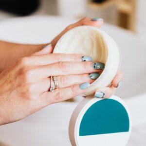 Close-up of hands applying skincare cream from a round container, showcasing elegant nail art.