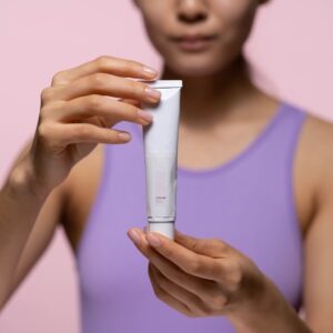 A woman in a purple tank top holds a skincare cream against a pink background in a studio setting.